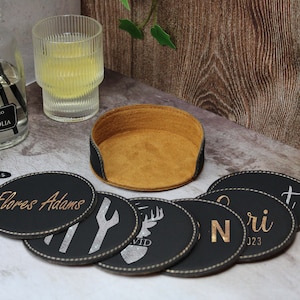 May include: Set of six black leather coasters with gold lettering and designs. The coasters feature names, a deer, a fork and knife, and a symbol. The coasters are arranged in a circle around a brown leather coaster holder.
