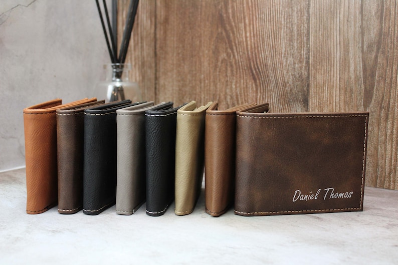 Puede incluir: Una fila de ocho carteras de cuero en varios colores, incluyendo marr&oacute;n, negro, gris y beige. La cartera de la derecha tiene el nombre "Daniel Thomas" grabado en blanco en la parte delantera.