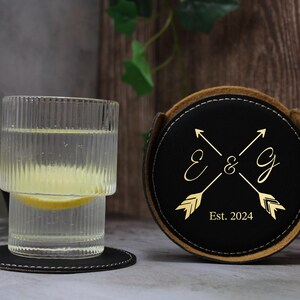 May include: A black leather coaster with a gold design of two arrows crossed with the initials "E & G" and the text "Est. 2024".
