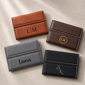 May include: Four rectangular card holders in tan, dark brown, gray, and black. Each features a fold-over flap with white contrast stitching. The tan holder displays L|M, the dark brown has CK in a circle, the gray says Laura, and the black has SK.