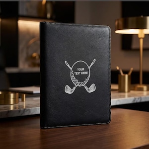 May include: Black leather golf score card holder with white stitching. The cover features a golf ball and crossed golf clubs design with the text "YOUR TEXT HERE" in the center. The holder is on a wooden surface.