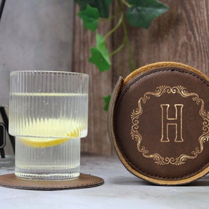 May include: A brown leather coaster with a gold monogram "H" in a decorative frame. The coaster is rolled up and sitting on a wooden surface next to a glass of water with a lemon slice.