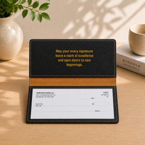 May include: A black leather checkbook cover with tan interior lining. The top flap features the gold text: May your every signature leave a mark of excellence and open doors to new beginnings. A blank check is inserted into the bottom slot.