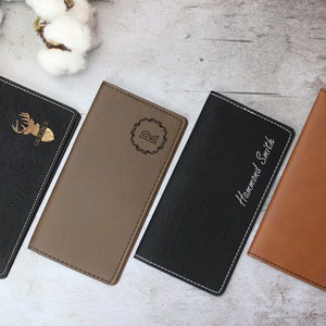May include: Four different personalized leather checkbook covers. The covers are black, brown, tan, and brown. The first cover has a deer silhouette with the word "Gurpack" underneath. The second cover has a monogrammed "R" in a circle. The third cover has the name "Hammond Smith" engraved on it. The fourth cover has a heart with an arrow through it.