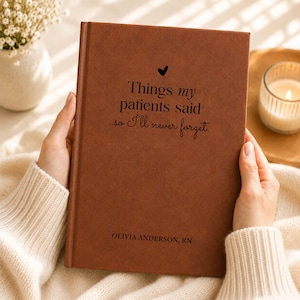 May include: A brown leather-like journal with a black heart icon and the text "Things my patients said ...so I'll never forget." and "OLIVIA ANDERSON, RN" printed on the front cover. The notebook has a smooth, matte finish and a vertical spine.