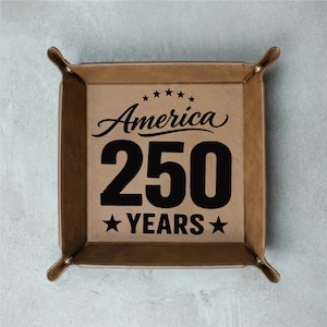 May include: A square, tan leather tray with raised edges and rounded corners. The center features the words "America 250 YEARS" in black, with stars above and below the number 250. The tray is designed to hold small items.