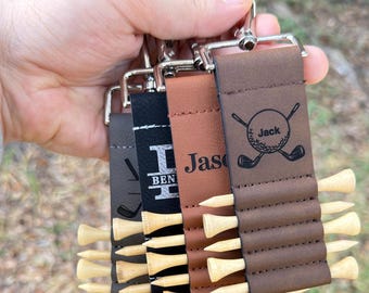May include: Four rectangular leather golf tee holders in gray, black, tan, and brown. Each holder features a silver metal clip and slots for wooden golf tees. The brown holder is engraved with the name Jack and a golf ball with crossed clubs.