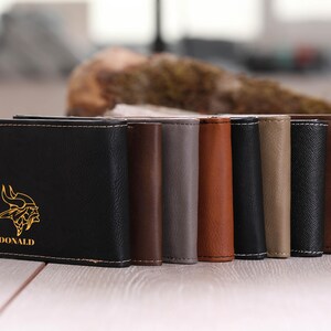 May include: A set of eight leather wallets in various colors, including black, brown, gray, and beige. The front of the black wallet features a gold-colored Viking helmet design and the name "DONALD" printed below it.