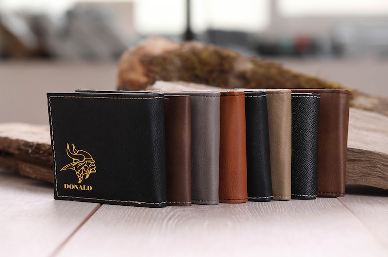 May include: A set of eight leather wallets in various colors, including black, brown, gray, and beige. The front of the black wallet features a gold-colored Viking helmet design and the name "DONALD" printed below it.