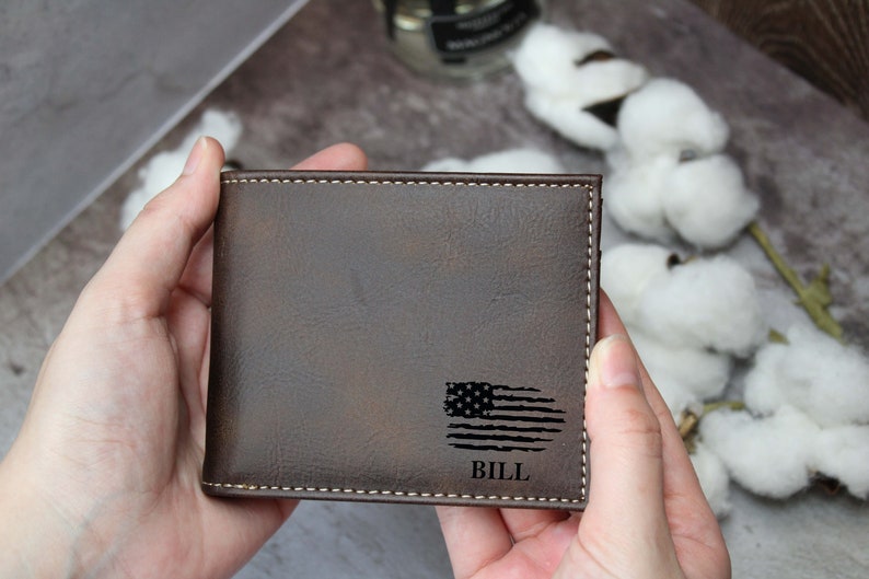 Puede incluir: Una cartera de cuero marr&oacute;n con un aspecto desgastado. Tiene un dise&ntilde;o de bandera estadounidense en blanco y negro y el nombre "BILL" grabado en ella.