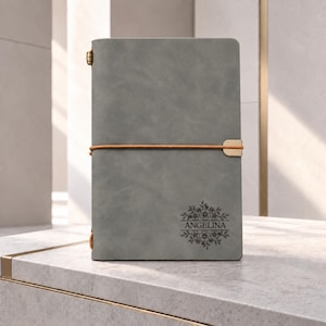 May include: A gray notebook featuring a tan elastic band closure and a gold metal accent. The cover is engraved with the name Angelina centered within a floral design consisting of small blossoms and leaves on the bottom right corner.