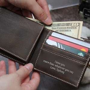 Puede incluir: Una cartera de cuero marr&oacute;n con un borde cosido blanco. La cartera est&aacute; abierta y tiene un billete de d&oacute;lar, tarjetas de cr&eacute;dito y un mensaje personalizado en el interior que dice "Te amo m&aacute;s que cualquier cosa que digas. Con amor, Sydney."