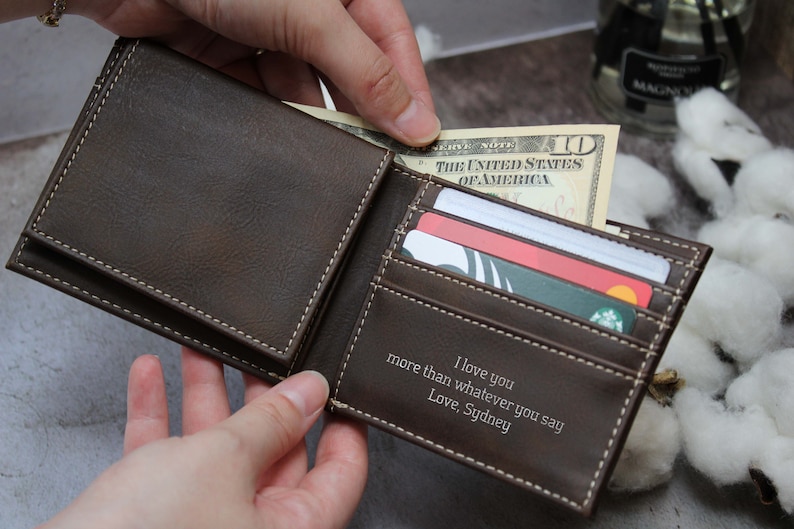Puede incluir: Una cartera de cuero marr&oacute;n con un borde cosido blanco. La cartera est&aacute; abierta y tiene un billete de d&oacute;lar, tarjetas de cr&eacute;dito y un mensaje personalizado en el interior que dice "Te amo m&aacute;s que cualquier cosa que digas. Con amor, Sydney."