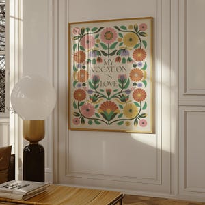 Saint Thérèse of Lisieux Catholic Wall Art Poster in Scandinavian Folk Floral Design with the phrase 'My vocation is love" centered. Around the text are folk-nspired flowers in hues of green, peach, salmon, pink, and yellow. The framed Catholic Quote Poster hangs on a living room wall.