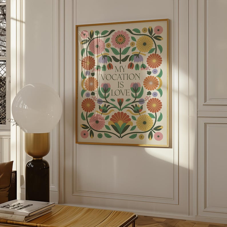 Saint Thérèse of Lisieux Catholic Wall Art Poster in Scandinavian Folk Floral Design with the phrase 'My vocation is love" centered. Around the text are folk-nspired flowers in hues of green, peach, salmon, pink, and yellow. The framed Catholic Quote Poster hangs on a living room wall.