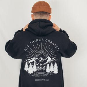 May include: Black hoodie with a white graphic on the back featuring mountains, trees, and the text "ALL THINGS CREATED" and "COLOSSIANS 1:16". The hoodie has a drawstring hood and long sleeves.