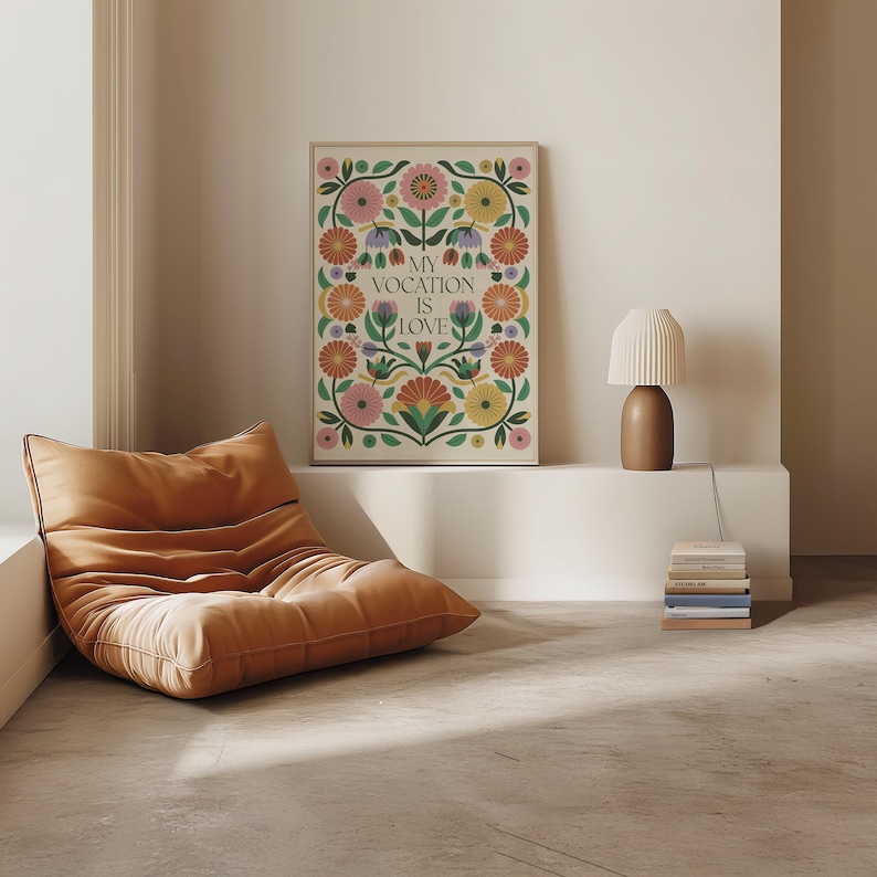 Saint Thérèse of Lisieux Catholic Wall Art Poster in Scandinavian Folk Floral Design with the phrase 'My vocation is love" centered. Around the text are folk-nspired flowers in hues of green, peach, salmon, pink, and yellow. The framed Catholic Quote Poster sits on a ledge in a mid-century modern living room.