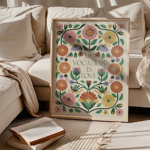 Saint Thérèse of Lisieux Catholic Wall Art Poster in Scandinavian Folk Floral Design with the phrase 'My vocation is love" centered. Around the text are folk-nspired flowers in hues of green, peach, salmon, pink, and yellow. The framed Catholic Quote Poster sits on the floor next to a beigh couch.