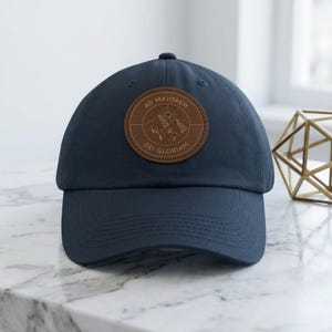 Pode incluir: Um boné de beisebol azul-marinho com um emblema circular castanho em imitação de pele na parte frontal. O emblema apresenta uma paisagem montanhosa gravada, um sol e o texto AD MAJOREM DEI GLORIAM numa borda circular central.