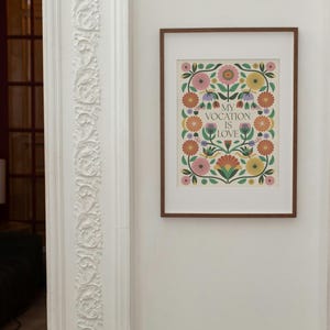 A framed art print featuring a floral design in shades of pink, orange, yellow, and green. The flowers are arranged around a central text that reads, 'My vocation is love.' The print has a vintage or folk-art style and is displayed on a white wall with ornate molding.