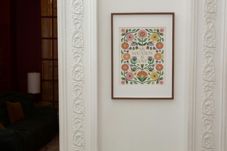 A framed art print featuring a floral design in shades of pink, orange, yellow, and green. The flowers are arranged around a central text that reads, 'My vocation is love.' The print has a vintage or folk-art style and is displayed on a white wall with ornate molding.