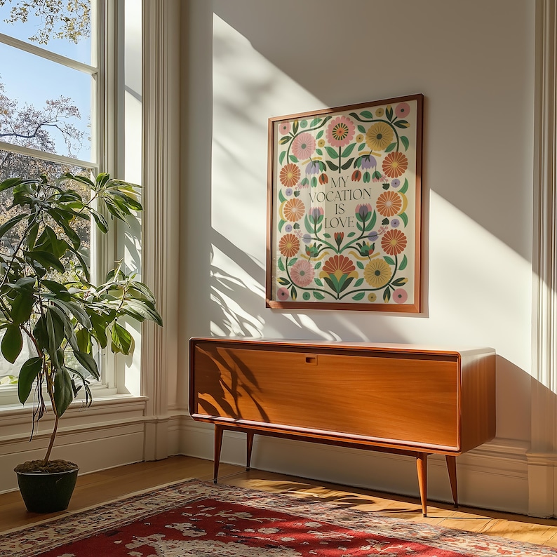 Saint Thérèse of Lisieux Catholic Wall Art Poster in Scandinavian Folk Floral Design with the phrase 'My vocation is love" centered. Around the text are folk-nspired flowers in hues of green, peach, salmon, pink, and yellow. The framed Catholic Quote Poster hangs on a wall in a mid-century modern living room.