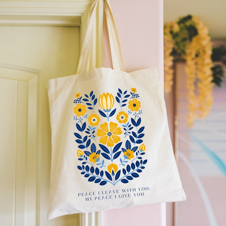 A natural colored Christian tote bag with blue and yellow floral design. The text on the bag reads 'Peace I leave with you; My peace I give you.'