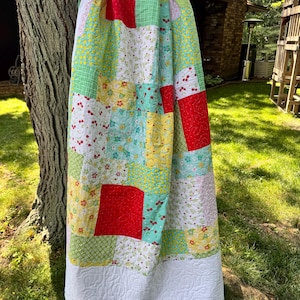 May include: A patchwork quilt featuring a variety of fabric squares in red, yellow, green, and white. The patterns include cherries, small flowers, and geometric shapes. The quilt is finished with a solid white border featuring a quilted swirl design.
