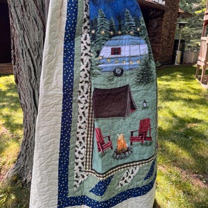 May include: A quilted blanket featuring a camping scene with a vintage camper, a brown tent, two red chairs, and a campfire. The border includes navy blue fabric with yellow dots and a white fabric with pinecone patterns, finished with cream backing.