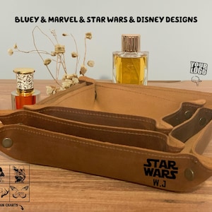 May include: A brown leather tray with three compartments. The tray has the words "Star Wars" and "W.J." engraved on the front. The tray is sitting on a wooden surface with a bottle of perfume and a bouquet of dried flowers.