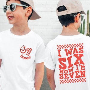 May include: White t-shirt with red graphics. The front features a number 67 with a checkered pattern and the name "Isaac". The back says "I WAS SIX NOW I'M SEVEN" in red, with a checkered border. The shirt is for a child.