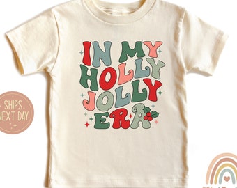 May include: Cream-colored short-sleeved t-shirt with the text "IN MY HOLLY JOLLY ERA" in a retro font. The text is in shades of red, green, and pink, with star accents and a holly sprig graphic. A rainbow logo is visible.