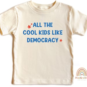 May include: Cream-colored t-shirt with the text "ALL THE COOL KIDS LIKE DEMOCRACY" in blue, with red star accents. The shirt is a short-sleeved design, suitable for children. A rainbow logo is visible in the bottom right corner.