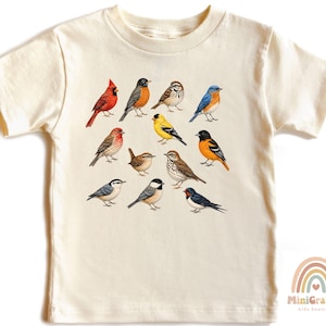 May include: A cream-colored short-sleeve t-shirt featuring a graphic print of twelve various songbirds in realistic colors, including a red cardinal, bluebird, and goldfinch, arranged in a grid pattern across the center of the chest.