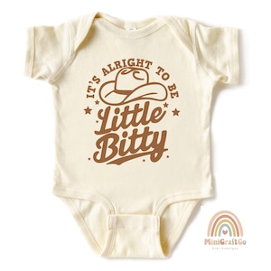 May include: A cream-colored baby onesie with a brown graphic that says "It's alright to be little bitty" with a cowboy hat graphic above the text.