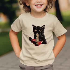 May include: A child wearing a cream colored t-shirt with a black cat eating ramen noodles from a red bowl. The cat is wearing a red collar.