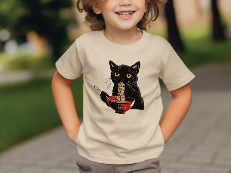 May include: A child wearing a cream colored t-shirt with a black cat eating ramen noodles from a red bowl. The cat is wearing a red collar.