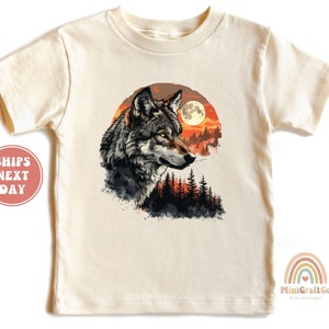 May include: A beige short-sleeved t-shirt with a graphic print of a wolf's head in front of a full moon and a forest. The wolf is looking to the right. The background is a watercolor-style sunset.