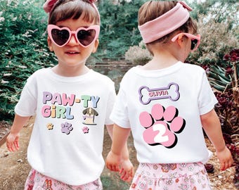 Könnte beinhalten: Zwei Kinder tragen weiße T-Shirts mit bunten Pfotenabdruck-Designs. Das vordere Shirt sagt "PAW-TY GIRL" und das hintere Shirt sagt "OLIVIA" auf einem Knochen und hat einen rosa Pfotenabdruck mit der Nummer "2" darin.