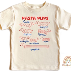 May include: A cream-colored short-sleeve t-shirt featuring red line art illustrations of dachshunds shaped like various pasta types. The text PASTA PUPS is printed in red above the dogs, with blue labels for fusilli, lasagna, farfalle, rigatoni, conchiglie, cavatappi, orzo, and spaghetti.
