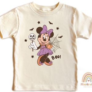 K&ouml;nnte beinhalten: Ein cremefarbenes Kurzarm-T-Shirt mit einer Grafik einer Zeichentrickmaus in einem lila Kleid mit Schleife. Die Maus h&auml;lt einen Totenkopf-Lutscher und einen K&uuml;rbis neben einem Spinnennetz. Das Design enth&auml;lt schwarze Flederm&auml;use, Sterne und BOO!