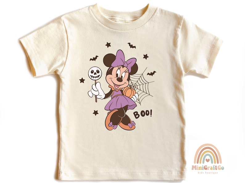 K&ouml;nnte beinhalten: Ein cremefarbenes Kurzarm-T-Shirt mit einer Grafik einer Zeichentrickmaus in einem lila Kleid mit Schleife. Die Maus h&auml;lt einen Totenkopf-Lutscher und einen K&uuml;rbis neben einem Spinnennetz. Das Design enth&auml;lt schwarze Flederm&auml;use, Sterne und BOO!