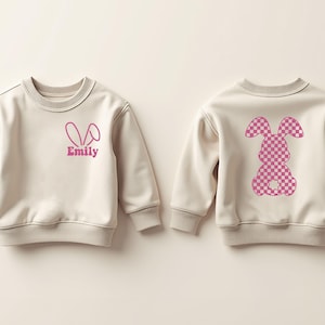 May include: Two cream-colored sweatshirts with pink bunny designs. One sweatshirt has a pink bunny with a checkered pattern on the back. The other sweatshirt has a pink bunny ear outline on the front with the name "Emily" underneath.