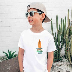 K&ouml;nnte beinhalten: Ein junges Kind tr&auml;gt ein wei&szlig;es T-Shirt mit einem Surfboard-Motiv und dem Text "Little Surfer Dude". Es tr&auml;gt au&szlig;erdem eine hellbraune Baseballkappe und eine blaue Sonnenbrille.