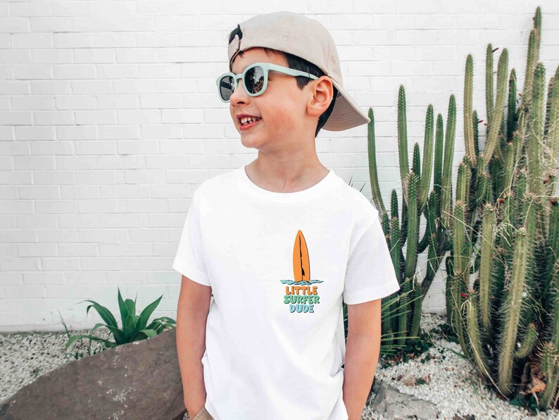 K&ouml;nnte beinhalten: Ein junges Kind tr&auml;gt ein wei&szlig;es T-Shirt mit einem Surfboard-Motiv und dem Text "Little Surfer Dude". Es tr&auml;gt au&szlig;erdem eine hellbraune Baseballkappe und eine blaue Sonnenbrille.
