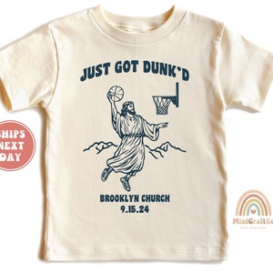 May include: A cream colored t-shirt with a graphic of a person dunking a basketball with the text "JUST GOT DUNK'D" and "BROOKLYN CHURCH 9.15.24".