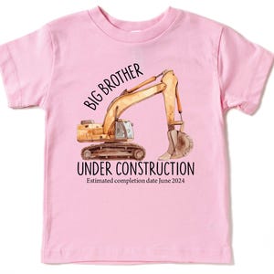 Pode incluir: Camiseta rosa claro para crian&ccedil;a com uma ilustra&ccedil;&atilde;o de uma escavadeira de constru&ccedil;&atilde;o. O texto diz "BIG BROTHER UNDER CONSTRUCTION" com uma data de conclus&atilde;o estimada em junho de 2024.