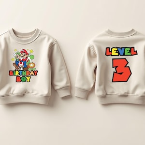 May include: Two beige sweatshirts with a video game theme. One sweatshirt features a graphic of Mario, Luigi, and Yoshi with the text "Birthday Boy". The other sweatshirt has the text "Level 3" in colorful letters.