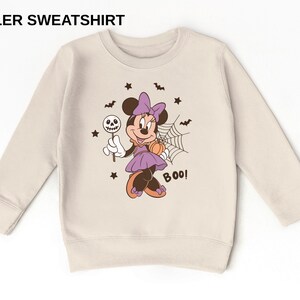 K&ouml;nnte beinhalten: Ein beiges Kleinkind-Sweatshirt mit dem Text TODDLER SWEATSHIRT oben. Die Grafik in der Mitte zeigt eine Cartoon-Maus in einem lila Kleid mit Schleife, die einen Totenkopf-Lutscher und einen K&uuml;rbis h&auml;lt, umgeben von Flederm&auml;usen, Sternen und einem Spinnennetz mit dem in Schwarz gedruckten Wort BOO!.