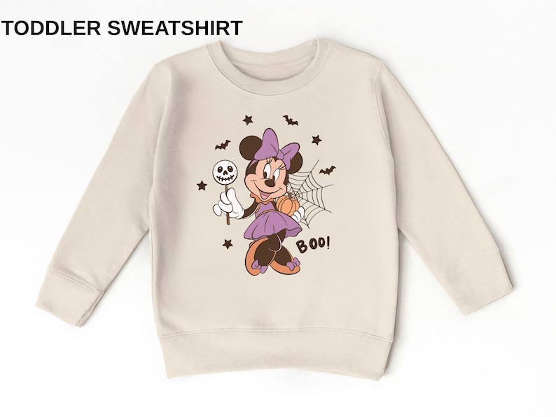 K&ouml;nnte beinhalten: Ein beiges Kleinkind-Sweatshirt mit dem Text TODDLER SWEATSHIRT oben. Die Grafik in der Mitte zeigt eine Cartoon-Maus in einem lila Kleid mit Schleife, die einen Totenkopf-Lutscher und einen K&uuml;rbis h&auml;lt, umgeben von Flederm&auml;usen, Sternen und einem Spinnennetz mit dem in Schwarz gedruckten Wort BOO!.